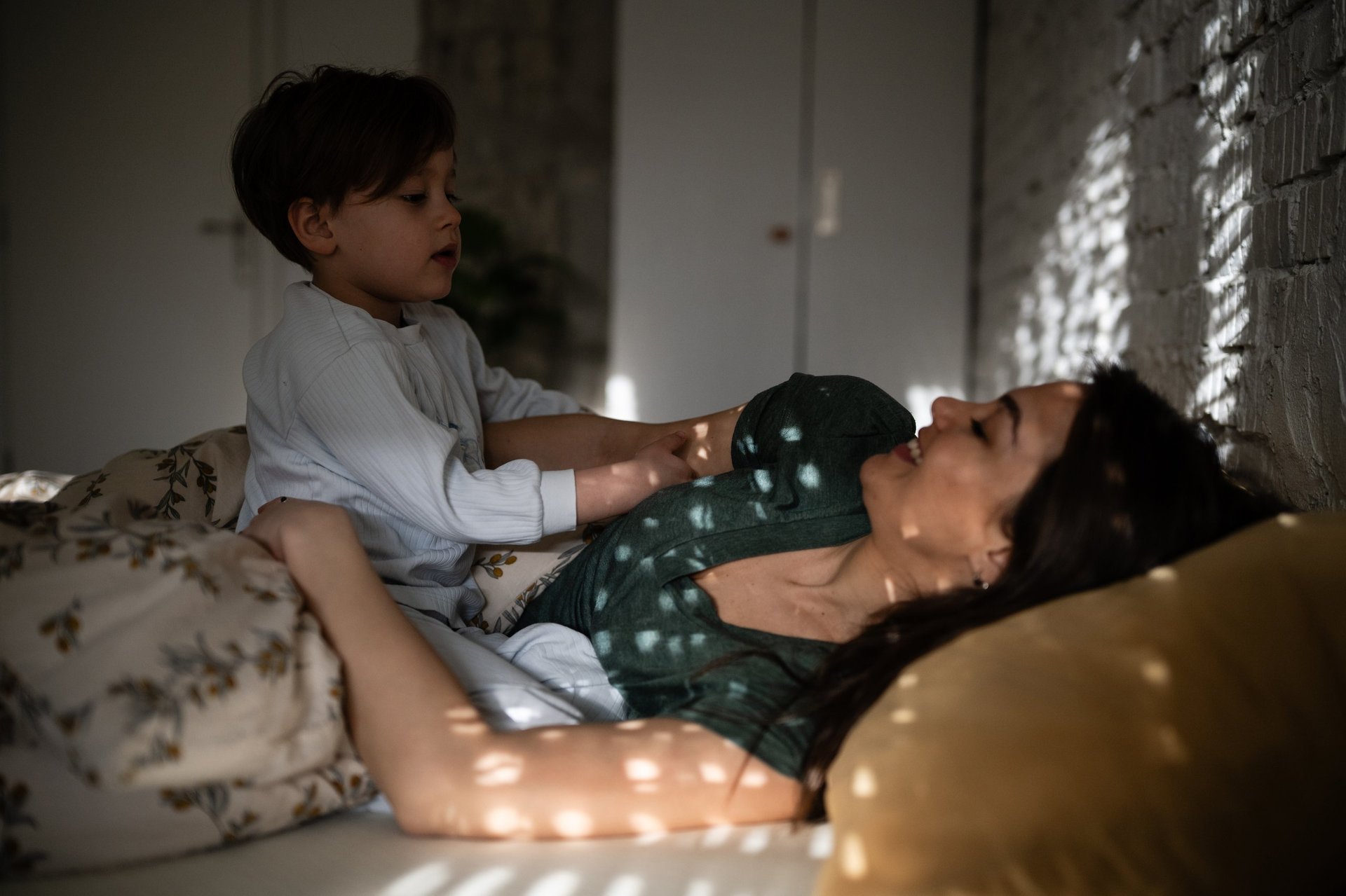Mom embracing young son in bed, both laughing together in sunlit bedroom, celebrating family love and happy parenting